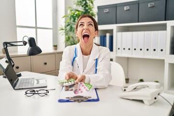 Young brunette doctor woman holding pills angry and mad screaming frustrated and furious, shouting with anger looking up.