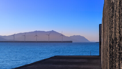 Vista de la silueta de montaña al horizonte sobre el mar con los molinos eólicos en el espigón al anochecer con cielo azulado