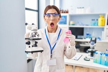 Young brunette woman working at scientist laboratory holding pink ribbon afraid and shocked with surprise and amazed expression, fear and excited face.