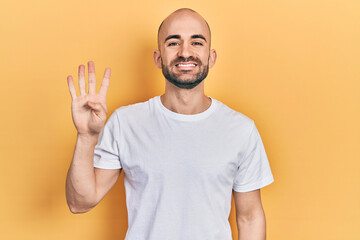 Young bald man wearing casual white t shirt showing and pointing up with fingers number four while smiling confident and happy.