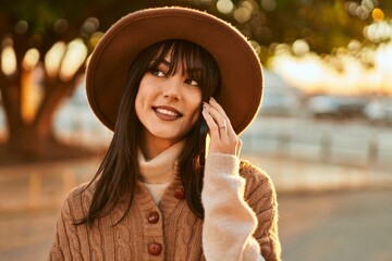 Brunette woman wearing winter hat smiling speaking on the phone outdoors at the city on sunset
