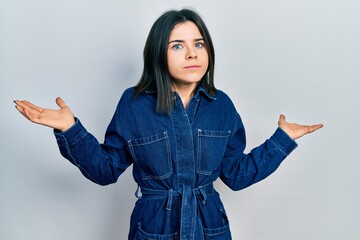 Young brunette woman wearing denim jumpsuit clueless and confused with open arms, no idea and...