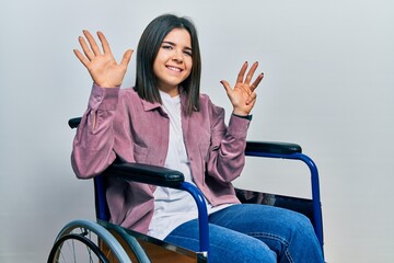 Young brunette woman sitting on wheelchair showing and pointing up with fingers number nine while...