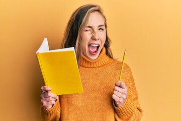 Young blonde woman holding book and pencil winking looking at the camera with sexy expression,...