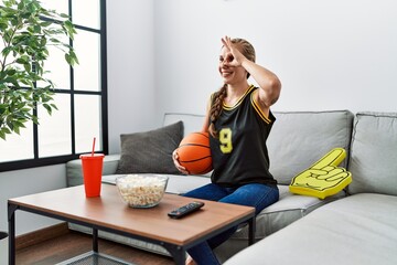 Young blonde woman holding basketball ball cheering tv game smiling happy doing ok sign with hand on eye looking through fingers