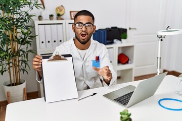 Young indian doctor showing blank clipboard scared and amazed with open mouth for surprise, disbelief face