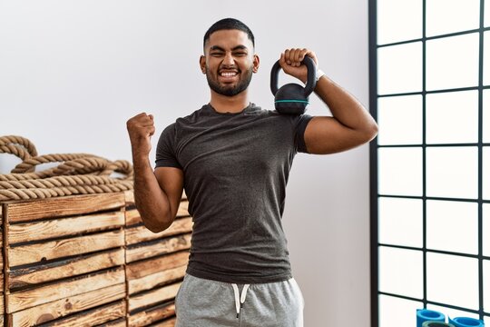 Young indian man wearing sportswear using dumbbells screaming proud, celebrating victory and success very excited with raised arm