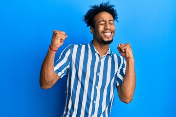 Young african american man with beard wearing casual striped shirt excited for success with arms...