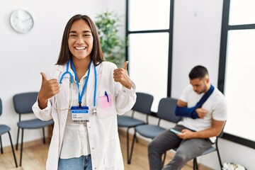 Young asian doctor woman at waiting room with a man with a broken arm success sign doing positive gesture with hand, thumbs up smiling and happy. cheerful expression and winner gesture.