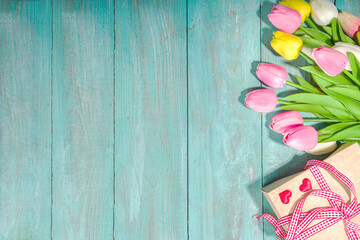 Spring holiday greeting card background with various color tulip flowers, gift box, flatlay on light blue high-colored wooden background top view copy space