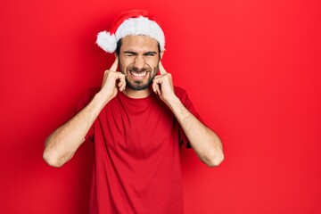 Young hispanic man wearing christmas hat covering ears with fingers with annoyed expression for the noise of loud music. deaf concept.