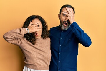 Middle age hispanic couple wearing casual clothes peeking in shock covering face and eyes with hand, looking through fingers with embarrassed expression.
