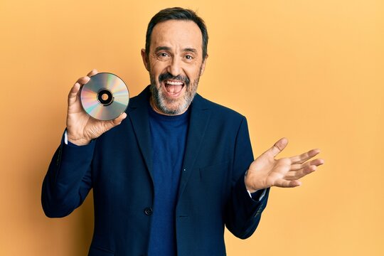 Middle age hispanic man holding compact disc celebrating achievement with happy smile and winner expression with raised hand