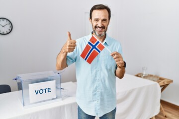 Middle age man with beard at political campaign election holding norway flag smiling happy and positive, thumb up doing excellent and approval sign