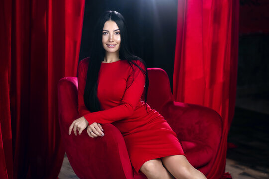 A beautiful brunette in a red dress sits in a red armchair in a dark apartment. Young woman in a sexy red dress sits in a chair and smiles seductively