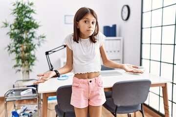 Young hispanic girl standing at pediatrician clinic clueless and confused with open arms, no idea concept.