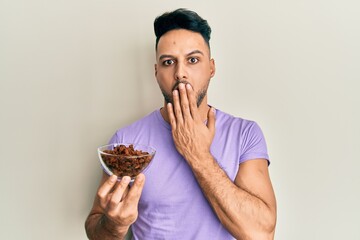 Young arab man holding raisins bowl covering mouth with hand, shocked and afraid for mistake. surprised expression - obrazy, fototapety, plakaty