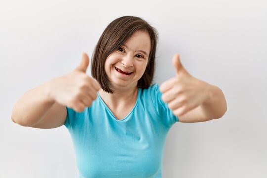 Young down syndrome woman standing over isolated background success sign doing positive gesture with hand, thumbs up smiling and happy. cheerful expression and winner gesture.