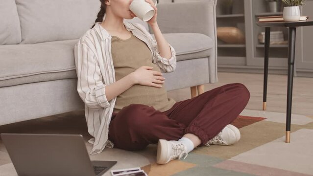 Cropped slowmo stab shot of young Caucasian pregnant woman drinking tea and stroking her belly sitting on floor at home with baby ultrasound scan and laptop lying nearby