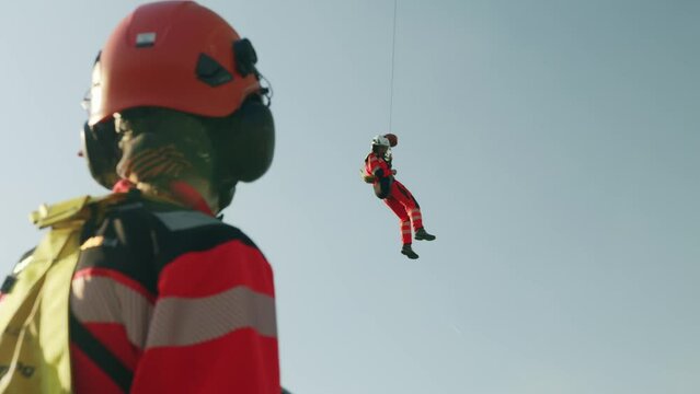 Rescuer hanging in the air from Helicopter hook and other one standing in foreground - sunny - slowmotion