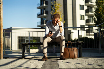 Young stylish businessman sitting on the beanch outdoors. Portrait of handsome man using the phone..