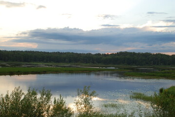 Sunset in the clouds over the lake. Above a small lake, along the edges of which a forest grows, a large white-gray cloud hangs, which hid the sun. The sun's rays shine through the cloud.