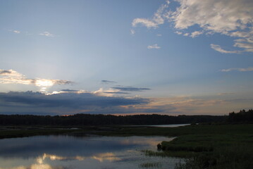 Sunset in the clouds over the lake. Above a small lake, along the edges of which a forest grows, a large white-gray cloud hangs, which hid the sun. The sun's rays shine through the cloud.