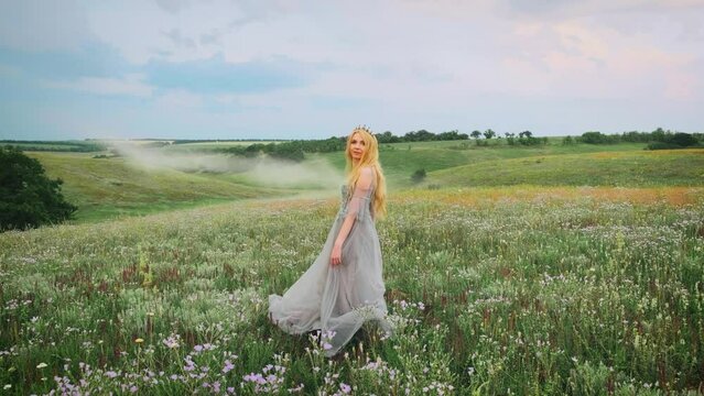 young happy blonde woman princess walks in green nature, whirls, dances fluttering skirt, silk blue gray dress waving in motion. Vintage style medieval summer clothes. candid girl bride. Crown on head