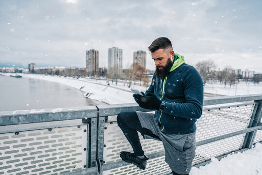 Handsome middle age man with a beard running and exercising outside on extremely cold and snowy day. Sport and fitness motivation theme. He using smart phone to track his activity data.