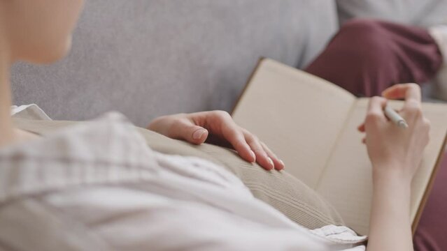 Medium closeup with slowmo of young pregnant woman writing in diary while resting on sofa at home stroking her belly