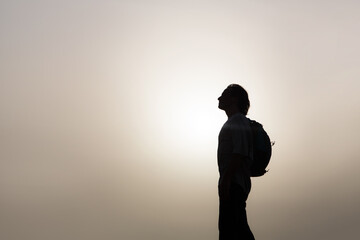 A male traveler with a backpack stands in profile against the background of the sky and sunset.