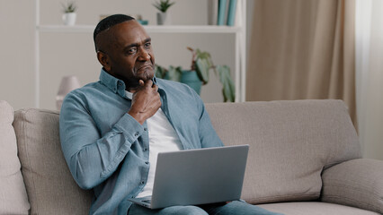 African american male freelancer sitting in room on sofa working remotely typing on laptop holds...
