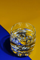 A transparent, glass small, rounded glass with clear water on a yellow and blue background separated by a clear border between them.