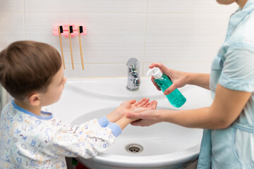 A young mother squeezes liquid soap from a dispenser onto her child's hands for washing hands at home over the washbasin in the bathroom. Personal hygiene. Selective focus. Close-up