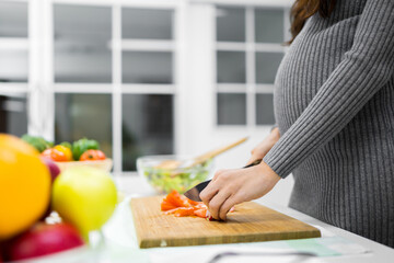 Obraz na płótnie Canvas Close-up photo of a healthy pregnant woman cooking healthy food, vegetable salad, at home kitchen. She is cutting red tomatoes.