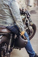 Naklejka premium Close up brave expirianced man driver wearing denim clothes and gloves sitting on a bike and holding a helmet in his hands. Close up motorcycle detail. Extreme concept