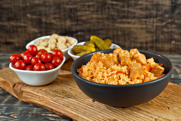 Pilaf with meat and carrots in a black bowl. Marinated mushrooms, tomato and cucumber on a black background. Food on a shabby table. Bowl on an old wooden board.