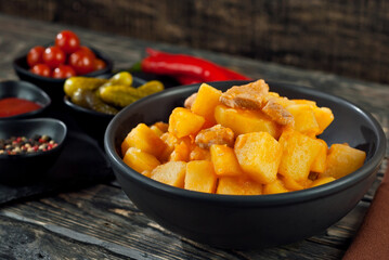 Boiled potatoes with meat in a black bowl. Seasonings and spices on a black wooden table. Ketchup, salt, pepper on an old shabby background.