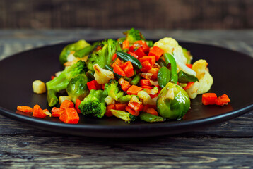 Mix of vegetables close up. Salad of various chopped vegetables. Roasted cabbage, carrots, broccoli, peas in a black plate. Food on the background of an old wooden board.