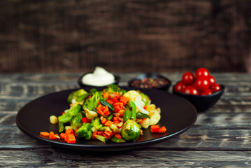 Mix of vegetables close up. Salad of various chopped vegetables. Roasted cabbage, carrots, broccoli, peas in a black plate. Food on a wooden board. Marinated tomatoes in the background.