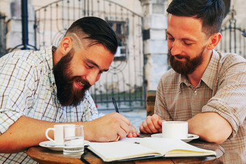 Two hipster businessmen working outdoors on the project, laughing and drinking coffee.