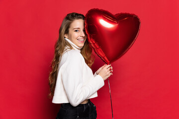 Beautiful smiling girl with long wavy silky hair, natural make up looking at camera with heart balloon isolated on red background. Valentines Day concept - obrazy, fototapety, plakaty