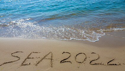 The inscription sea 2022 on the sand by the water and the rising wave, seashore beach vacation by the sea