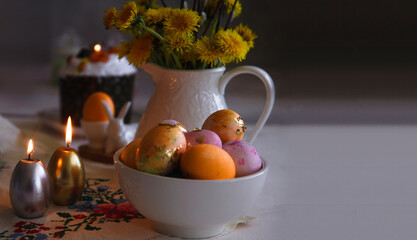 Easter still life with orange and pink eggs, burning candles, bouquet of yellow dandelions on white table and gray background, copy space