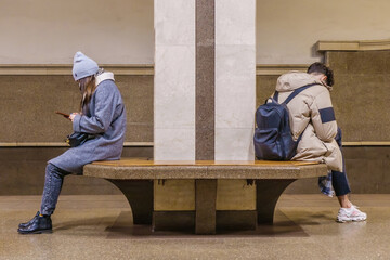 Two young people, a boy and a girl, are sitting in the lobby of a metro station with their backs to each other. Both of them are passionate about their phones, not paying attention to anything else.