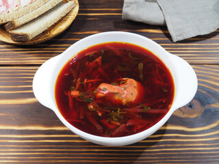 Delicious hot beetroot soup in a white bowl, bread with bacon on a wooden table, napkin, top view, flat layout. Traditional red borscht with meat and various vegetables for lunch