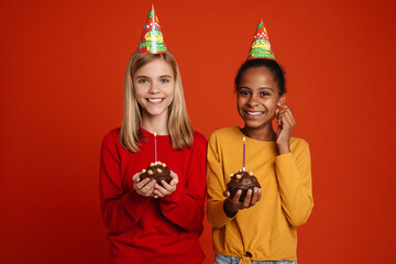Multiracial girls wearing party cones laughing while posing with cakes