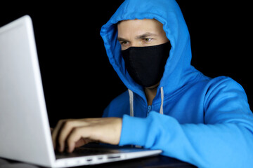Man in mask and blue hoodie sitting with laptop on black background. Concept of cyber crime, hacking and safety work during coronavirus - Powered by Adobe