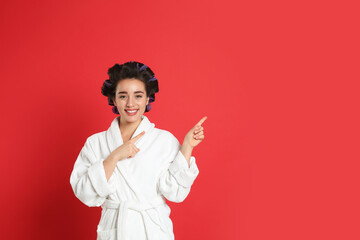 Happy young woman in bathrobe with hair curlers on red background, space for text