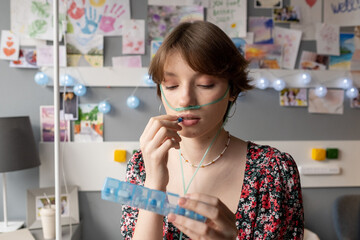 Teenage girl with oxygen nose pipe taking pills and looking at medicaments in long plastic...